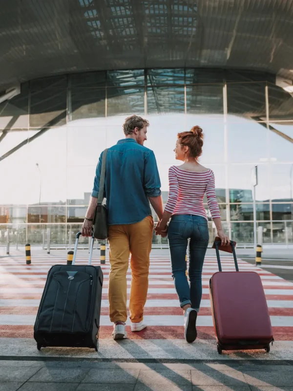 Beautiful loving couple with travel suitcases holding hands in airport Full length back view portrait of young man and his charming girlfriend walking and carrying their trolley bags. They looking at each other and smiling